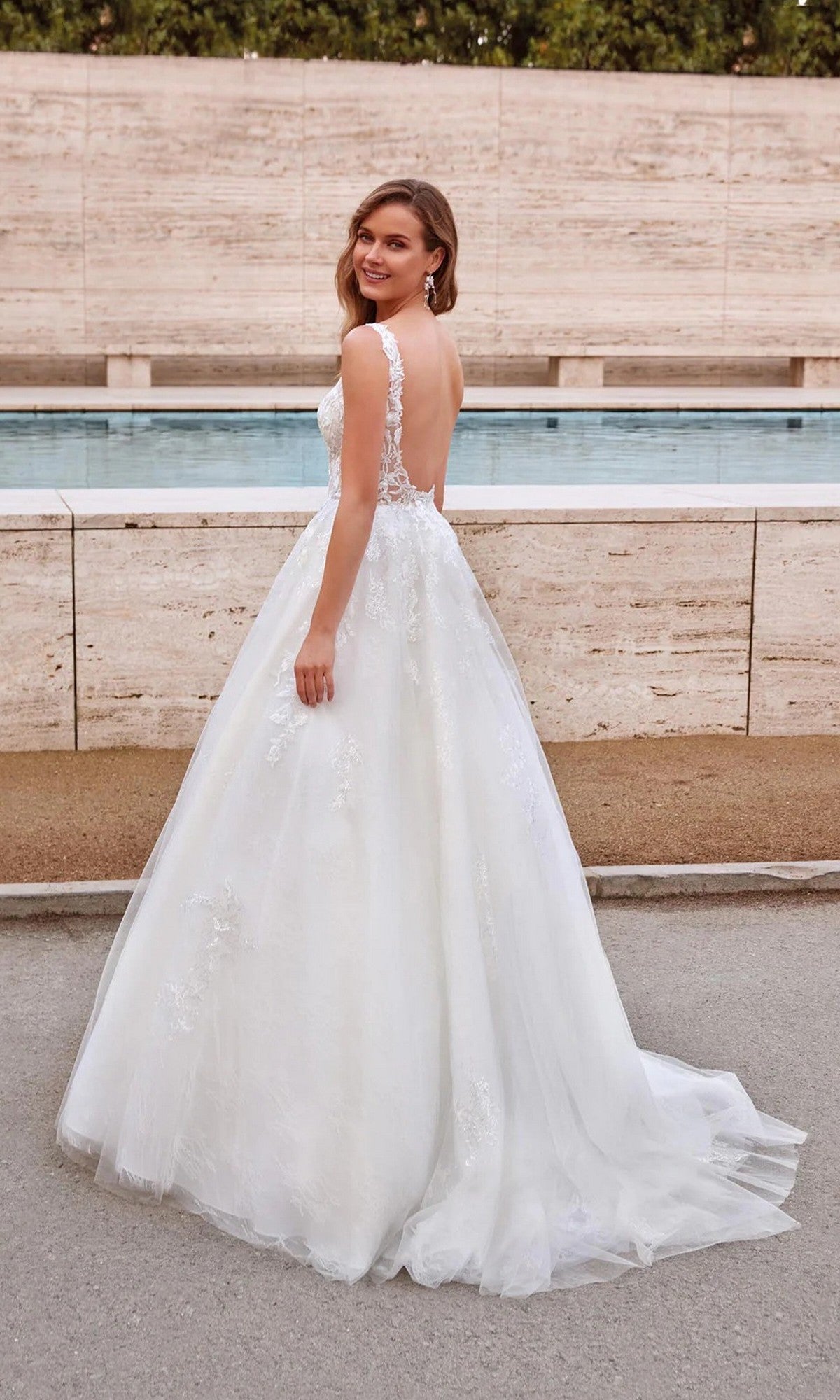 A woman in the Alyce 7039 long white dress, a sleeveless lace ball gown with a low back, stands outdoors and looks over her shoulder.